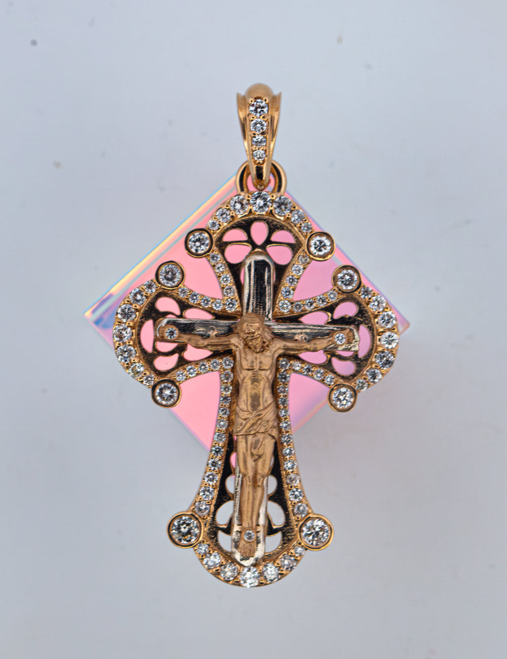 A 14k gold crucifix pendant with a central figure of Jesus and an ornate filigree cross adorned with small diamonds tones, shown against a light gray background with a pink iridescent card behind it.