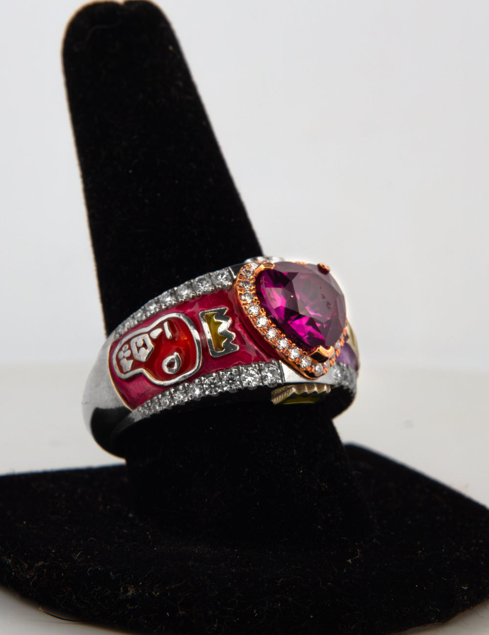 A custom enamel ring displayed on a black velvet ring stand. The ring features a silver band paved with small diamond stones, a red enamel section with a white and gold football helmet, and a large heart-shaped pink gemstone surrounded by rose gold  halo  and diamond stones.