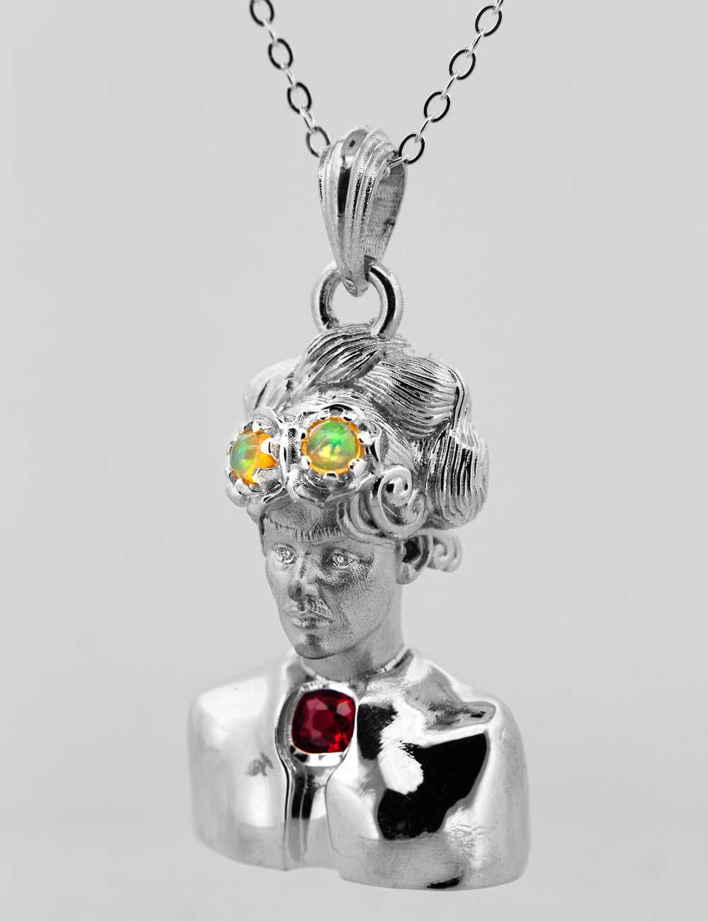 A silver pendant necklace featuring a Frida Kahlo bust with three colorful  opal gemstones in her hair and a red gemstone on her chest, shown against a plain light gray background.