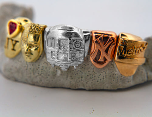 A close-up of a ring featuring five New York City subway system themed charms in gold, silver, and rose gold, displayed on a textured gray stone stand spiderman in 14k green gold. Yankees and MetroCard 