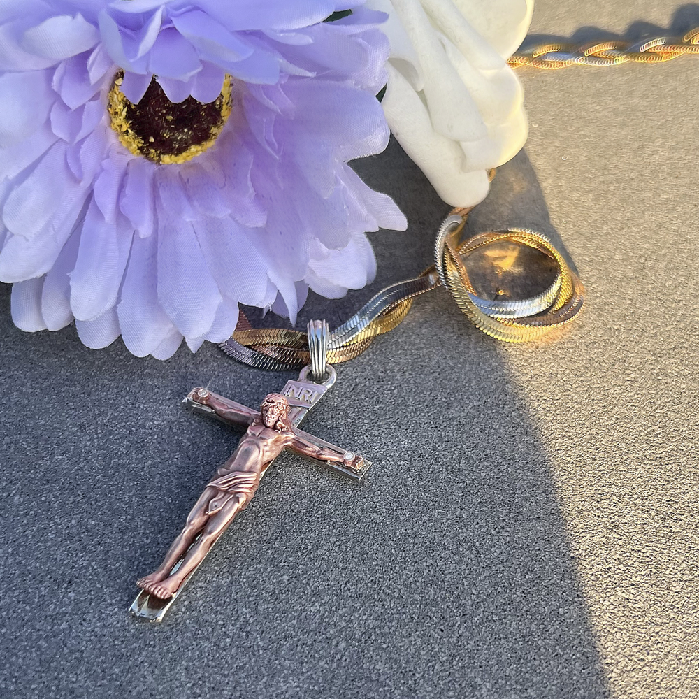 A rose gold crucifix pendant necklace with a matching chain, styled with a purple flower and a white rose on a textured gray surface.